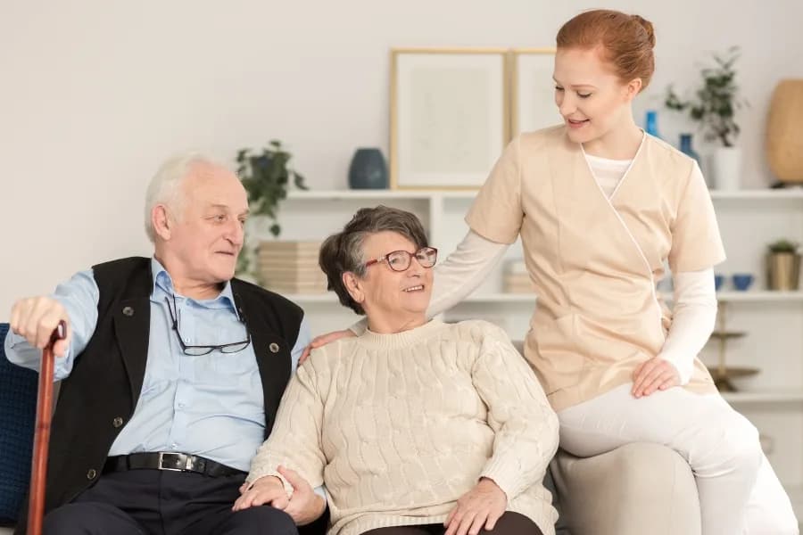 A young carer in uniform smiles with an elderly couple seated together in a bright living room.