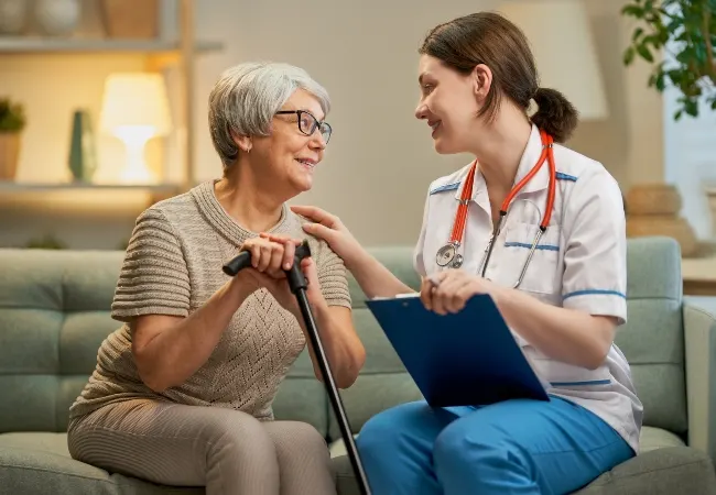 A smiling care worker with a stethoscope and clipboard sits chatting with an elderly woman holding a walking stick.