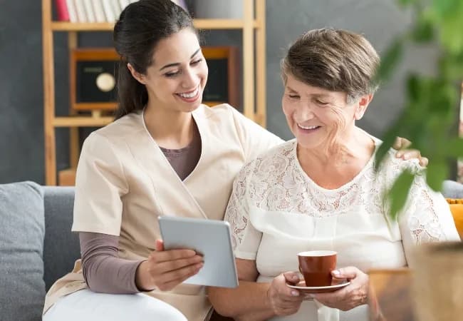 A carer and smiling older woman holding a cup of tea look at a tablet together on a sofa.