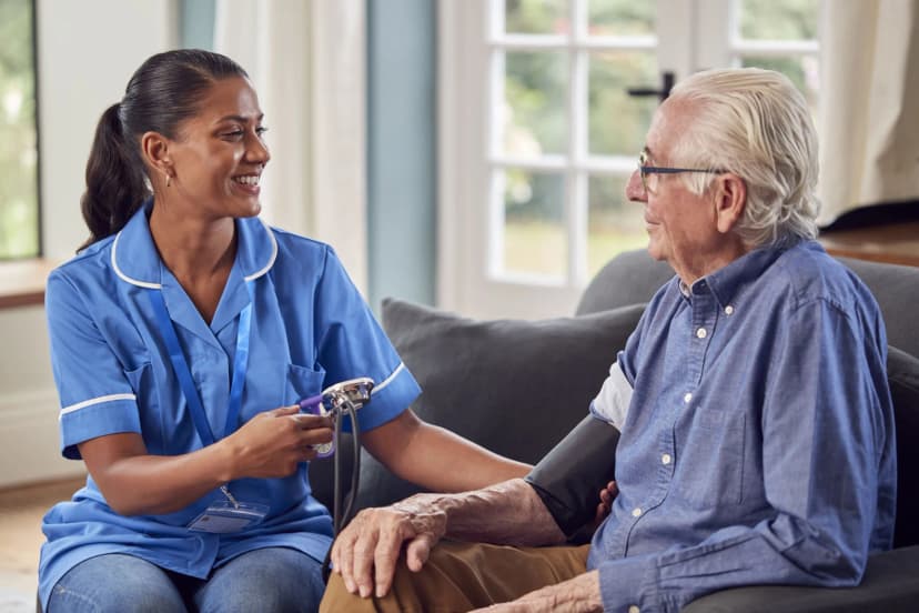 A smiling care worker in a blue uniform checks an elderly man's blood pressure as they sit together at home.