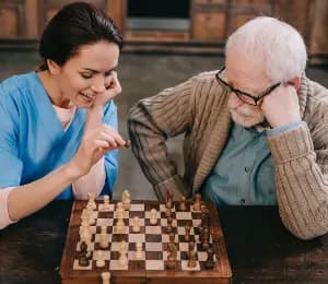 A smiling carer in blue plays chess at a table with an older man wearing glasses.